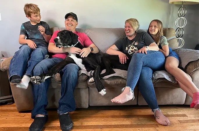 A family of 4 sits on a couch, smiling with a Great Dane laying across them.